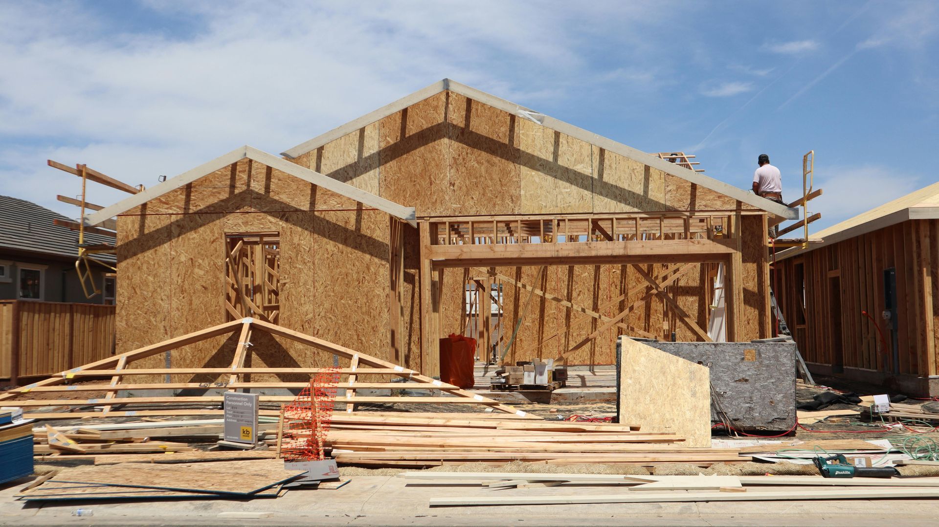 Wood-framed house under construction with scaffolding, lumber piles, and two workers on the roof