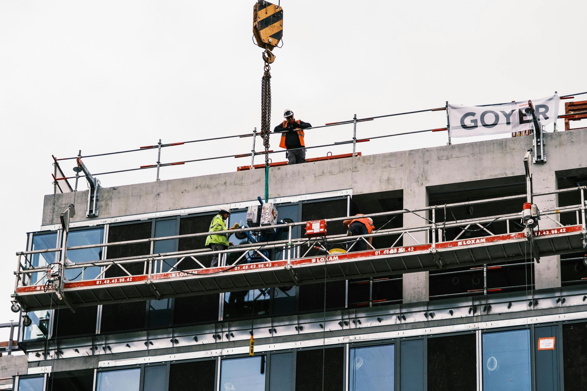 Construction workers on scaffolding atop a building, with a crane hook overhead.