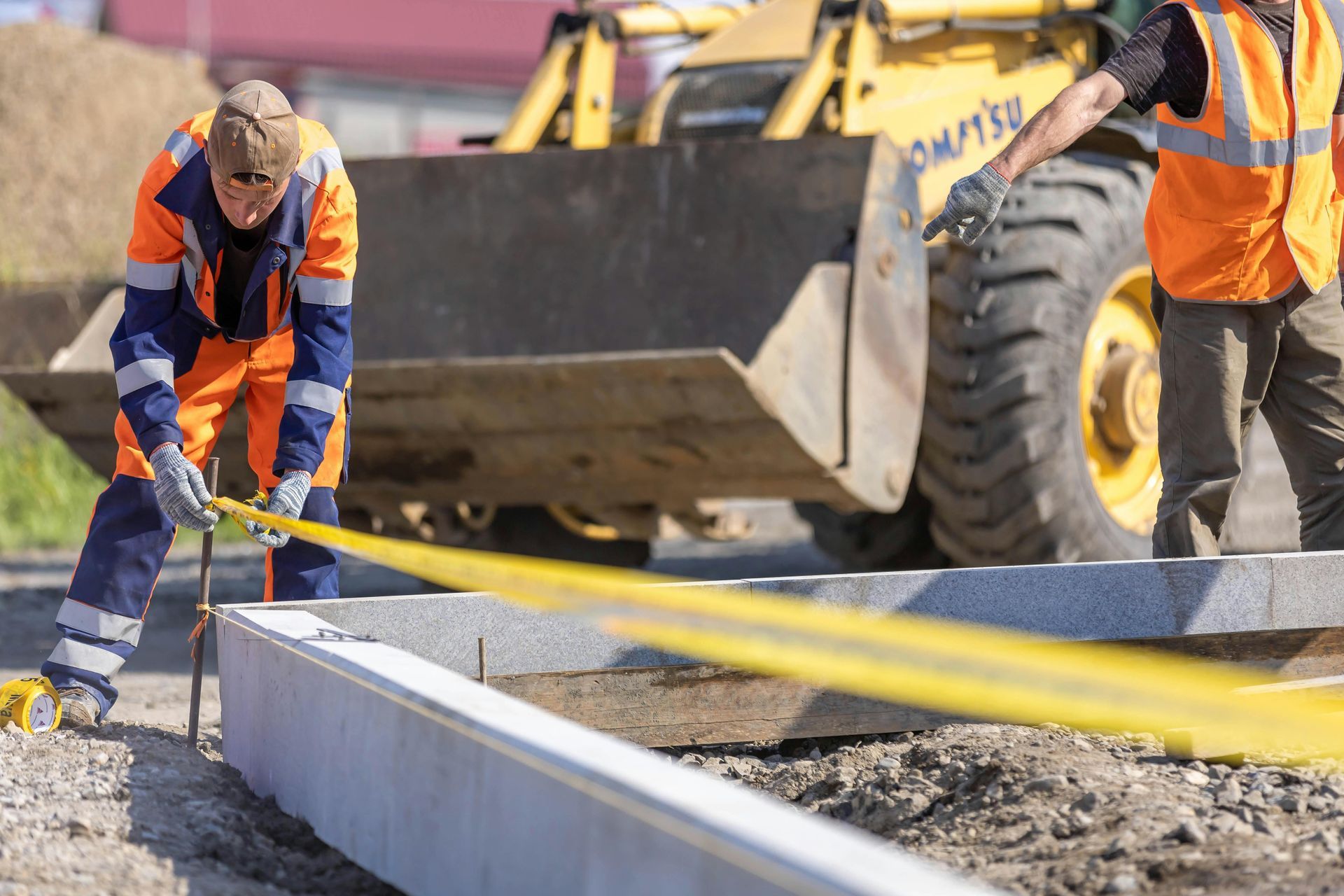 Construction workers pouring concrete into a curb form beside a yellow loader at a work site