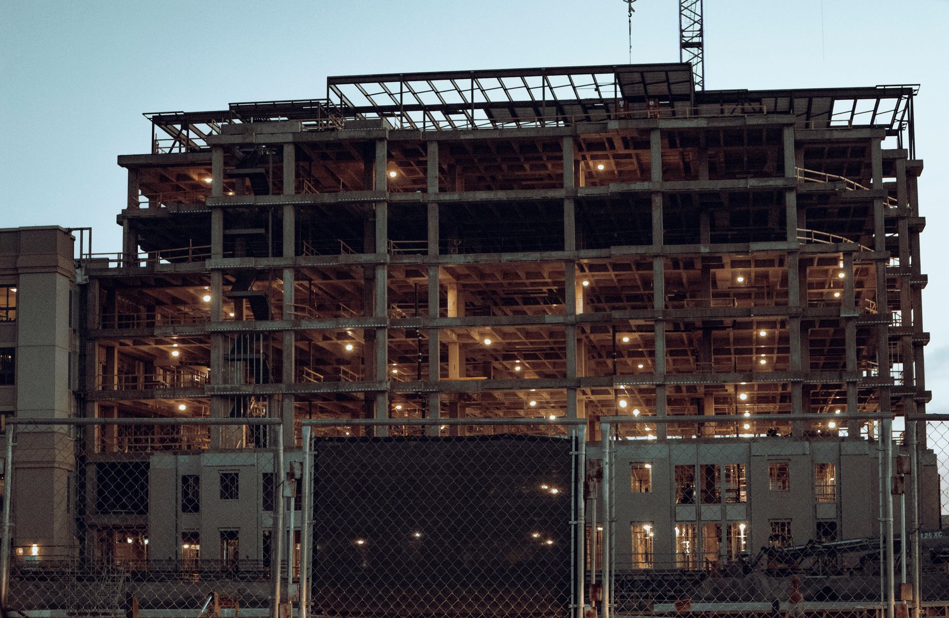 Partially built steel-framed building at dusk, lit with construction lights behind a chain-link fence