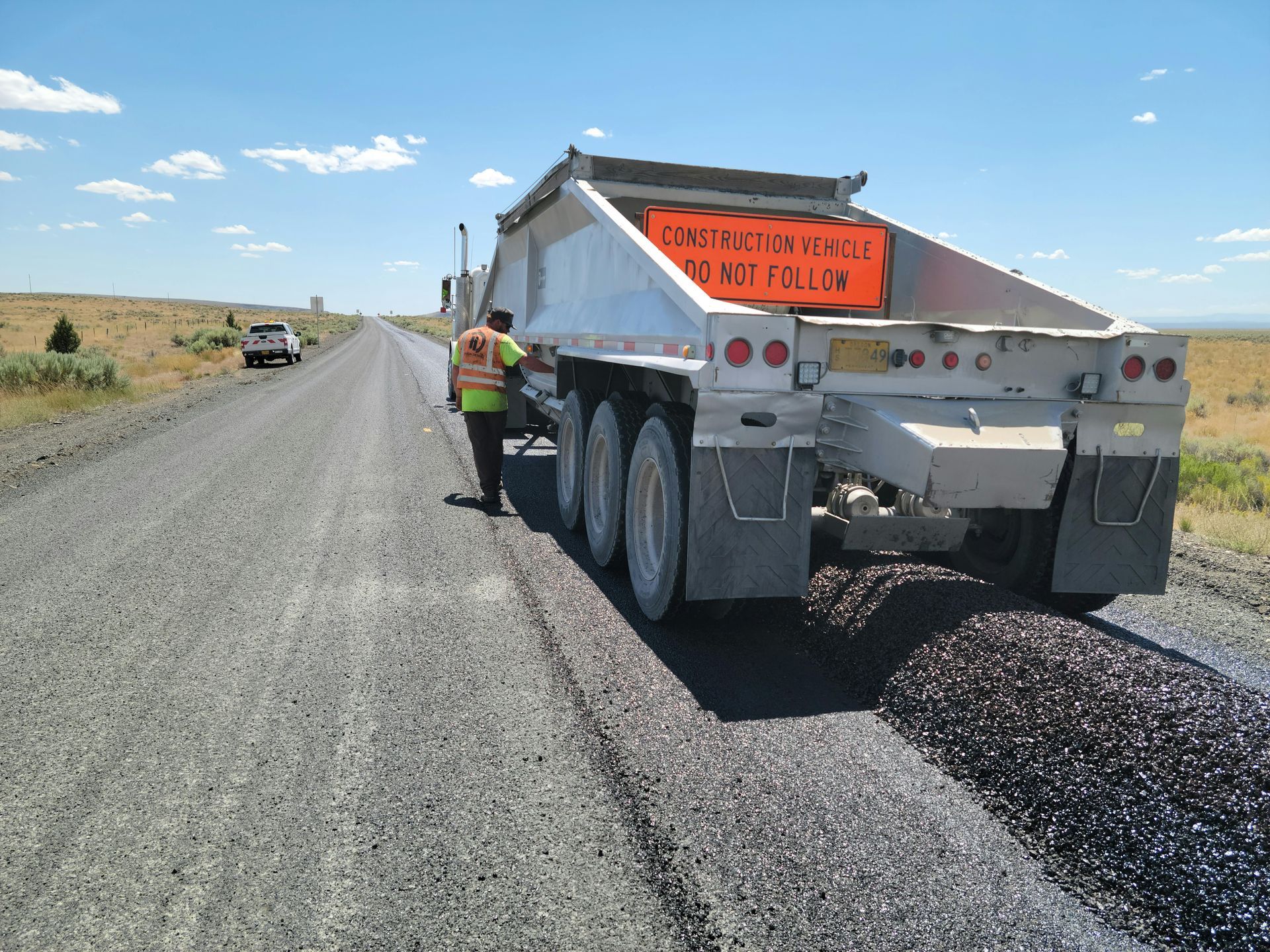 Road worker beside dump truck spreading gravel on a rural dirt road under a clear blue sky