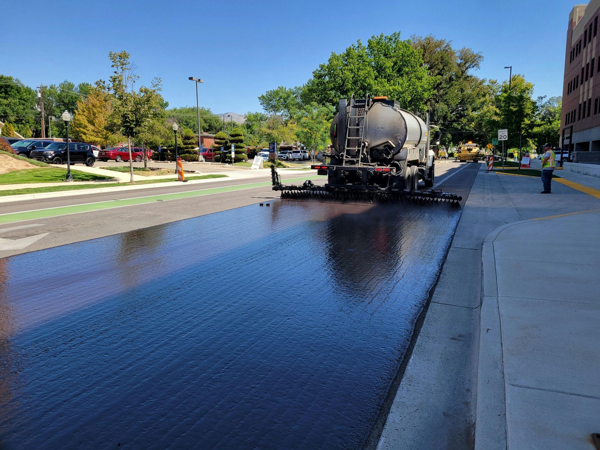 Road resurfacing truck spraying fresh black asphalt beside a sidewalk on a sunny street