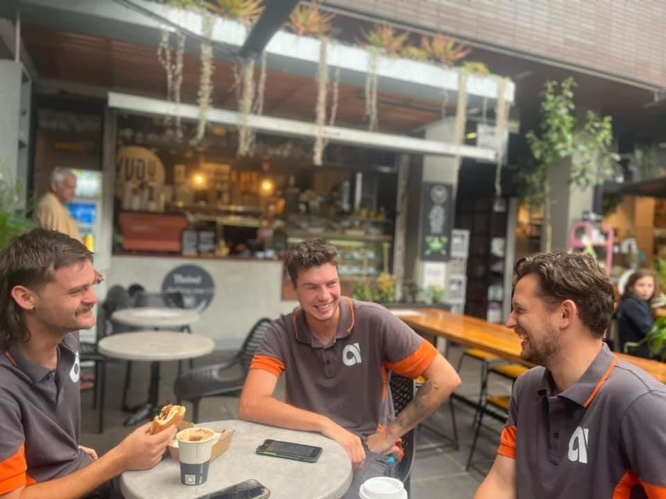 Three men in gray shirts with orange accents laugh at an outdoor cafe.