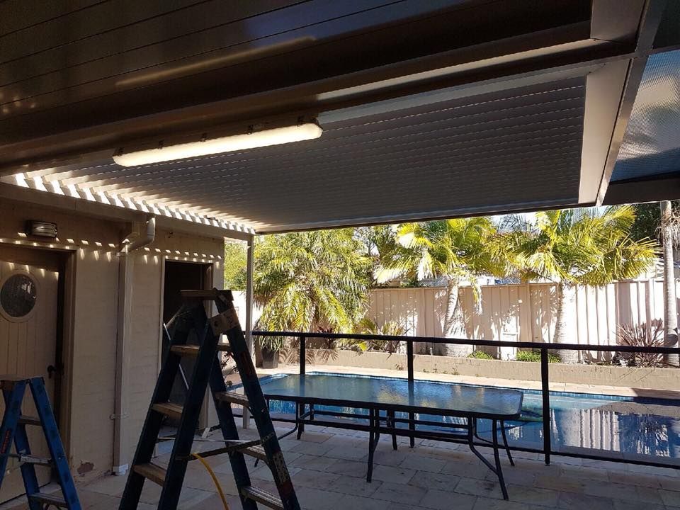 Patio with a metal roof, outdoor table, ladder, and string lights, overlooking a pool.