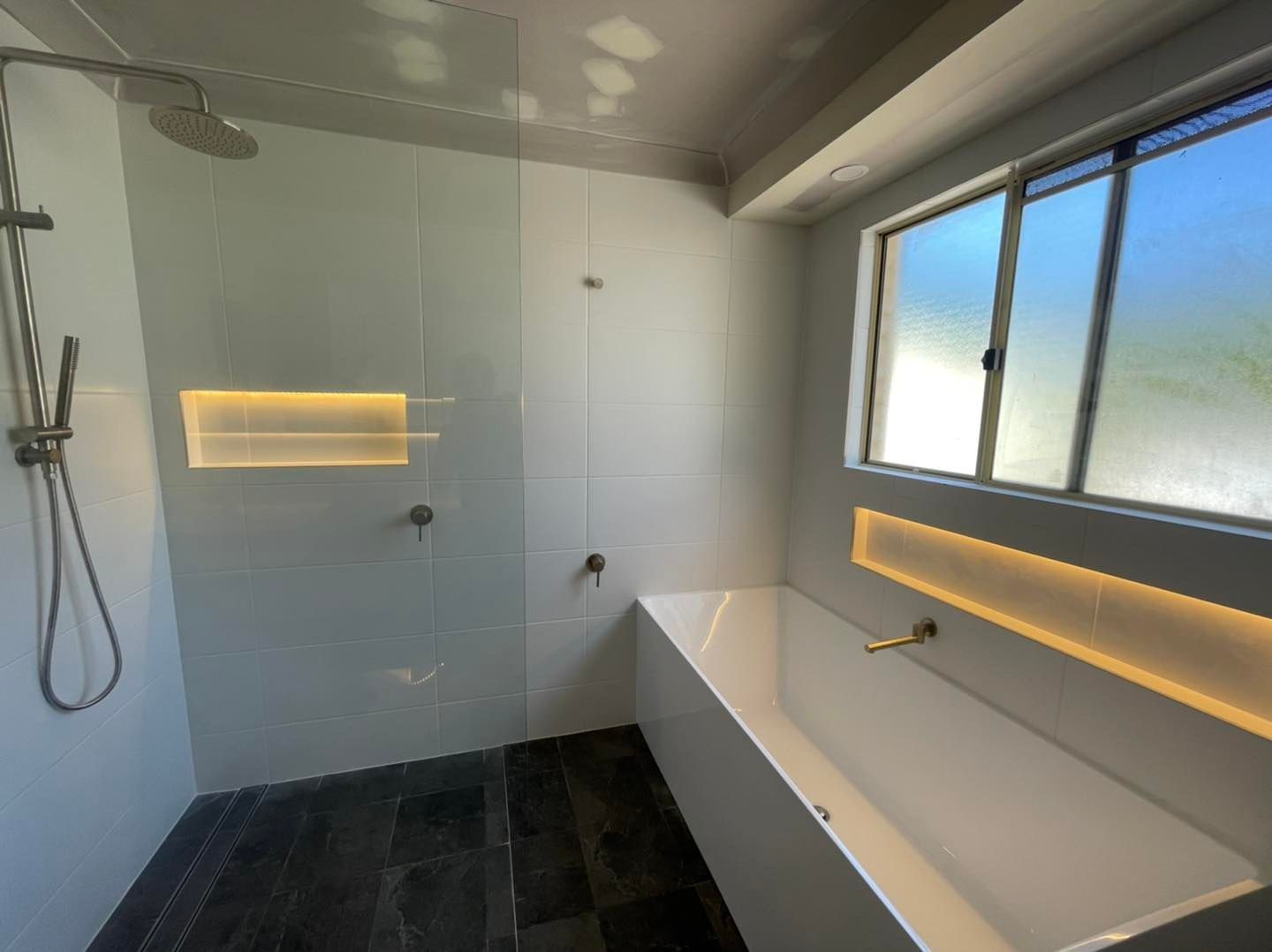 Modern bathroom with glass shower, bathtub, window, and recessed lighting. Black tile floor.