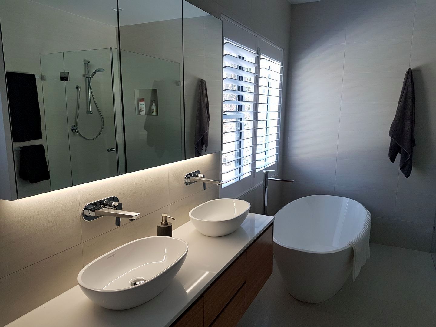 Modern bathroom with double sinks, bathtub, and mirrored wall. Gray walls, light wood accents, and window with shutters.