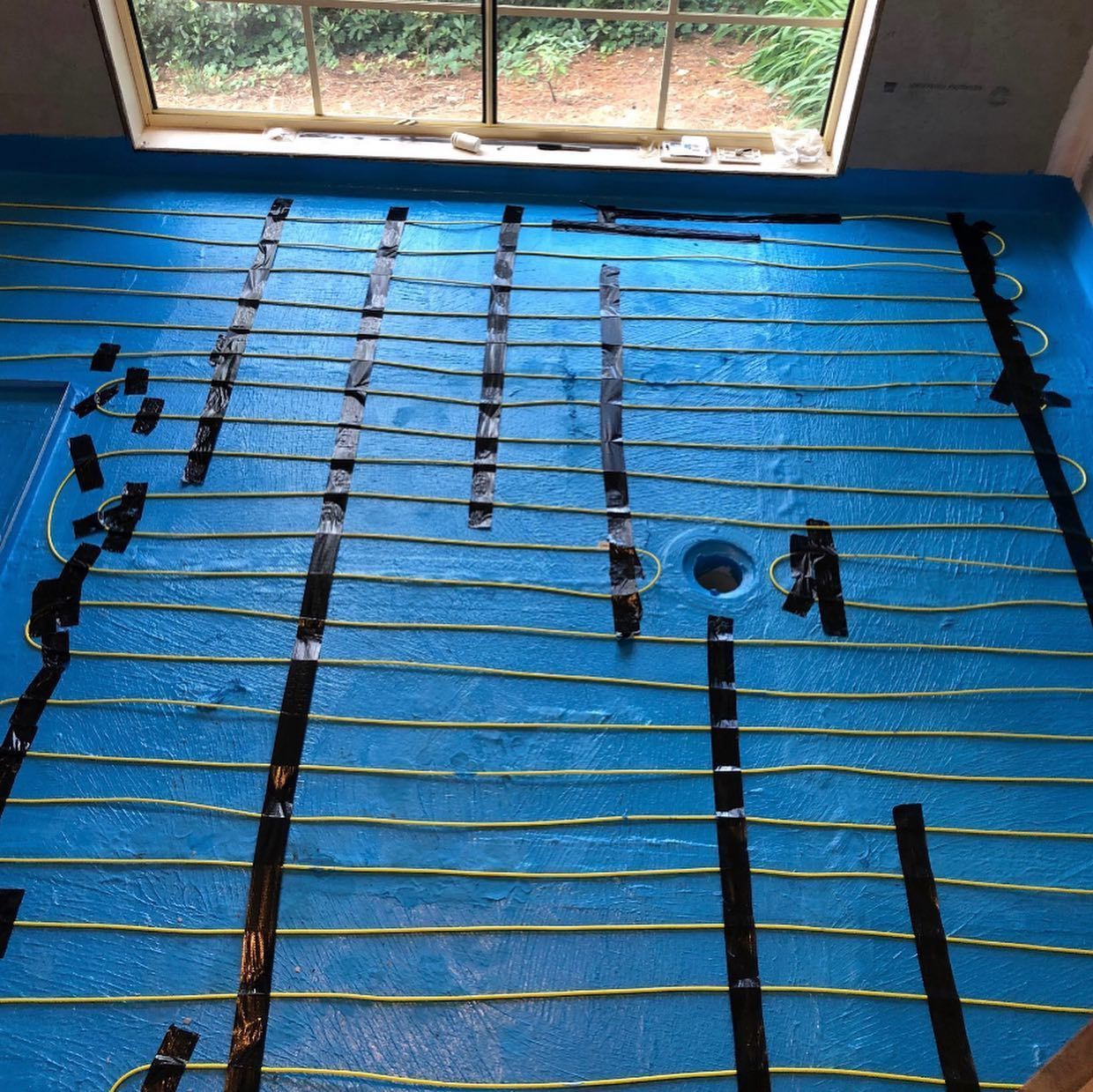 Blue bathroom floor with installed radiant heating cables taped down, near a window.