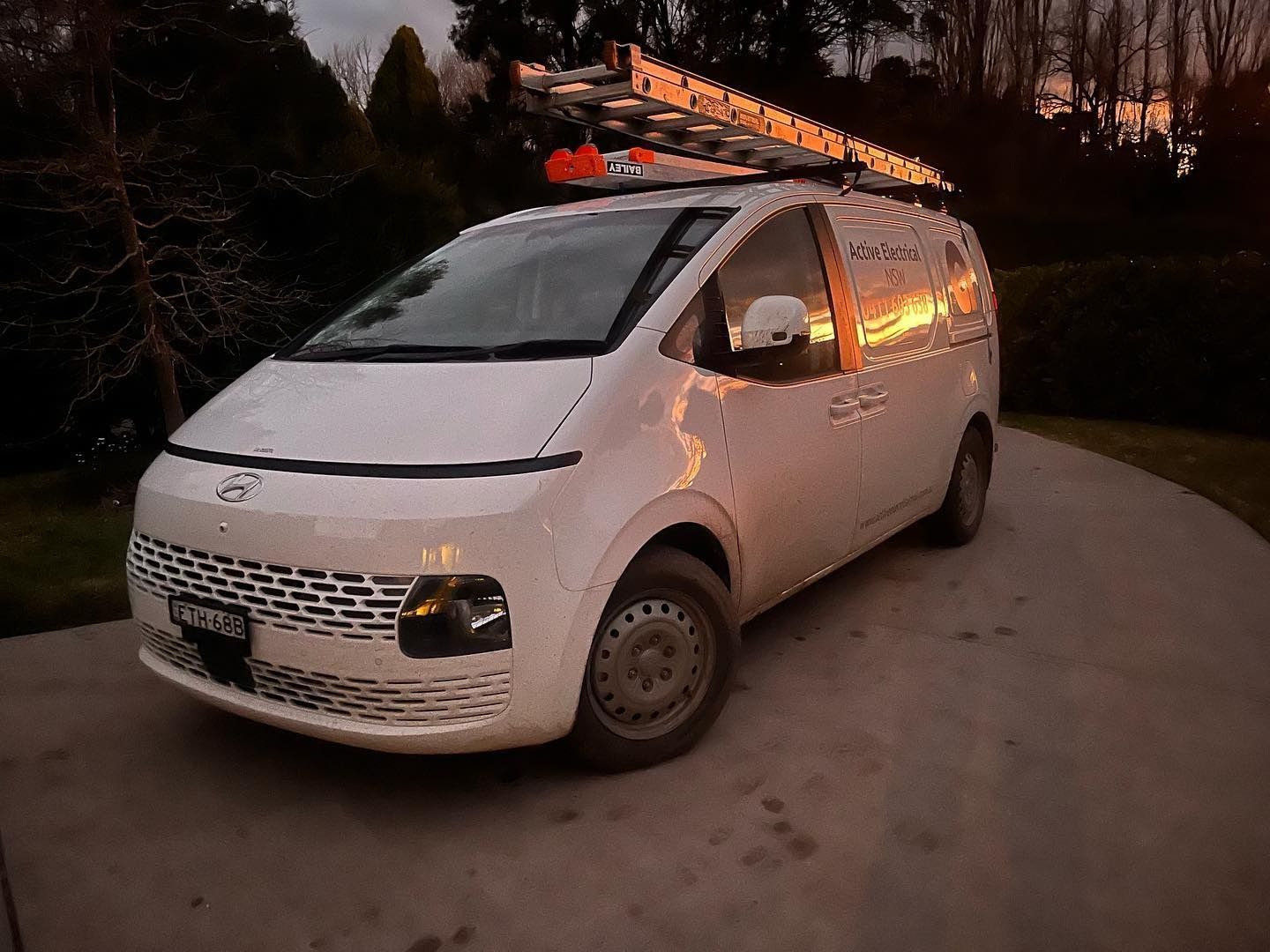 White van parked on a driveway at dusk; ladder on the roof, sunlight reflecting off the windows.