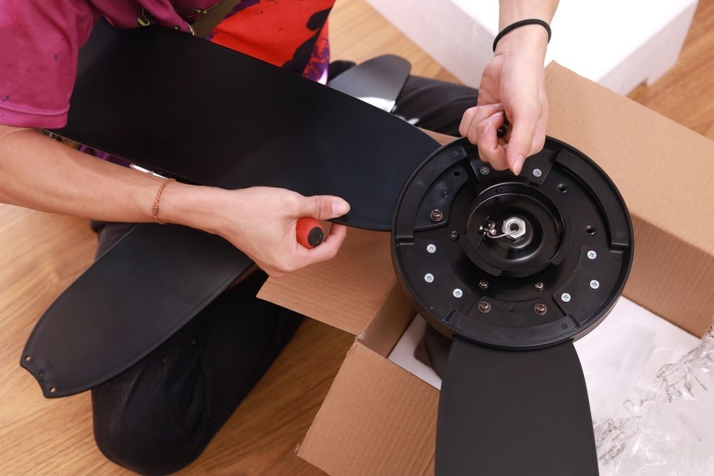 Person attaching a black fan blade to a ceiling fan motor. Cardboard box is visible.