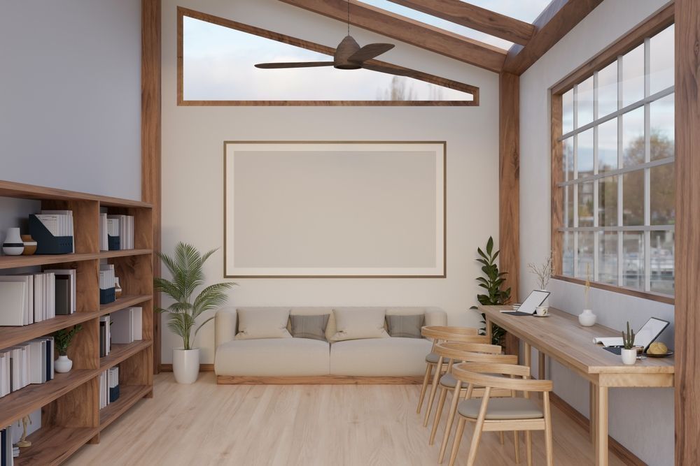 A light-filled room with wooden beams, bookshelves, a couch, desk, and large windows.