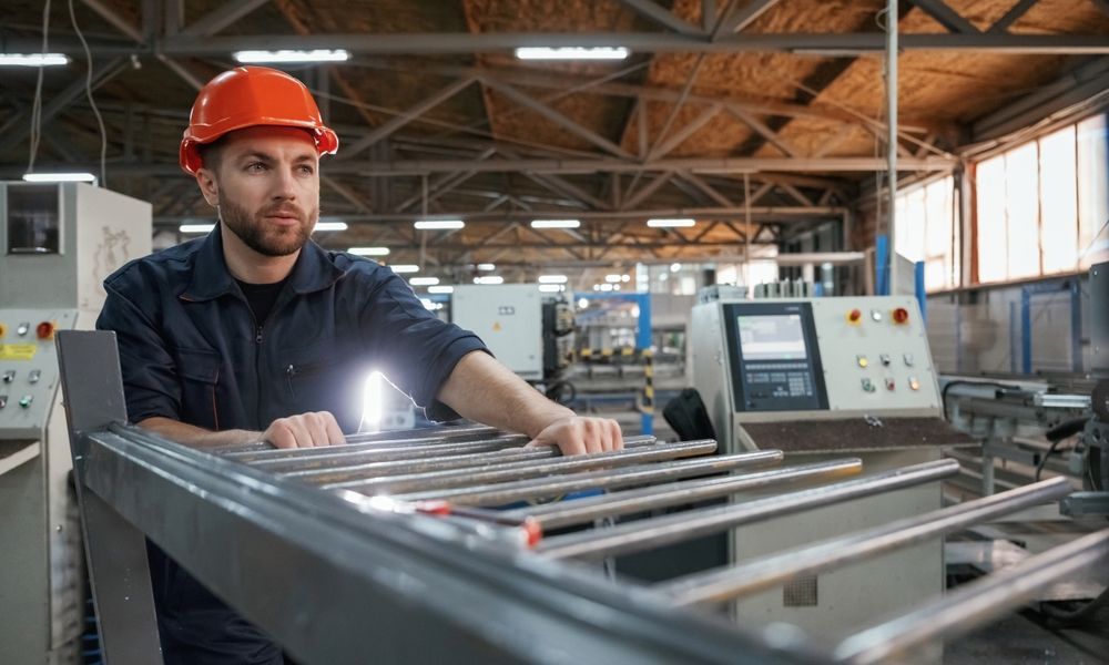Man in orange hard hat examines metal rods in a factory setting.