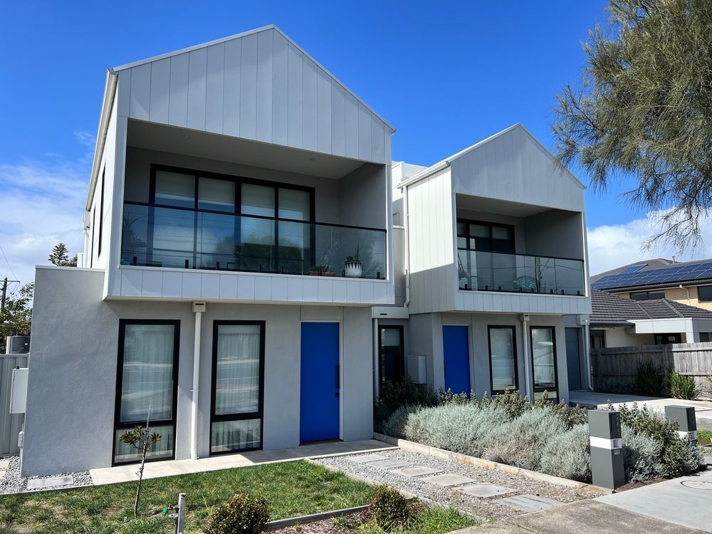 Two-story modern buildings with blue doors, black-framed windows, and balconies, under a bright blue sky.