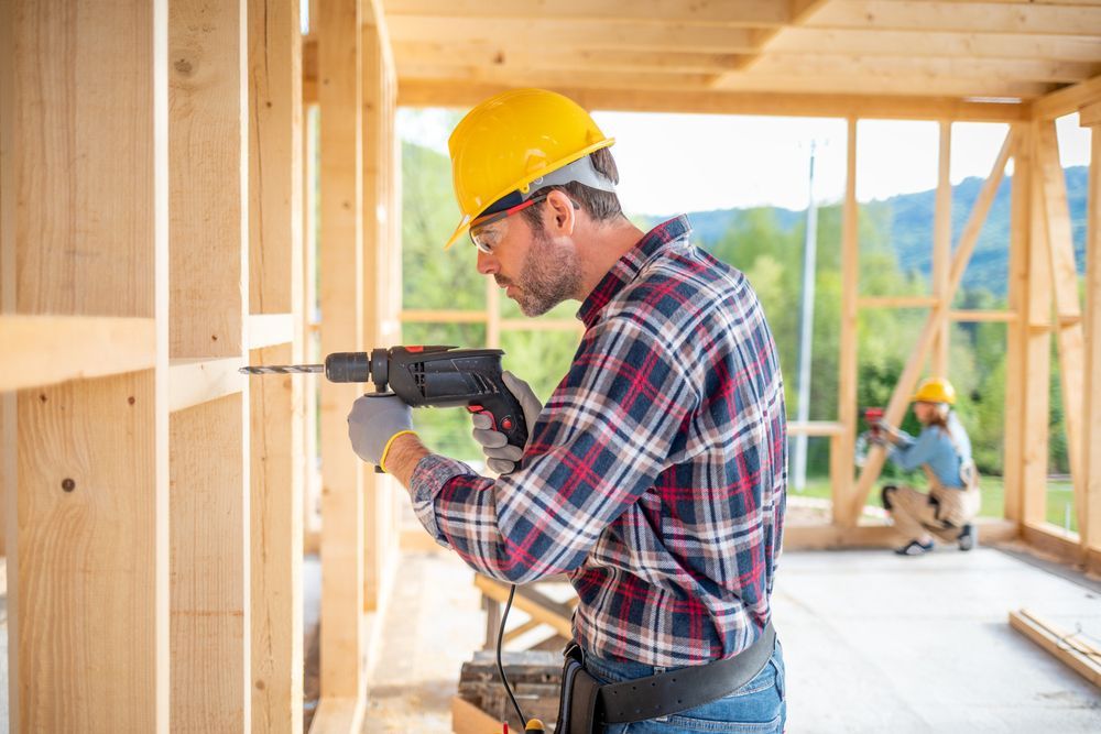 Construction worker using a drill on a wooden frame, wearing a hard hat and safety glasses.