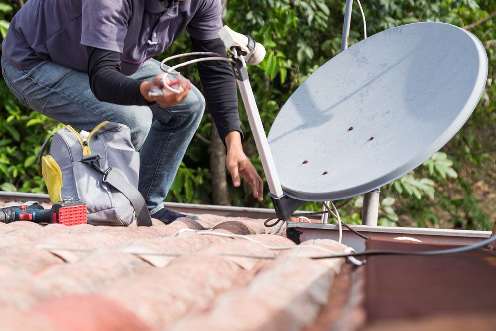 Person installing a satellite dish on a rooftop, with tools nearby.