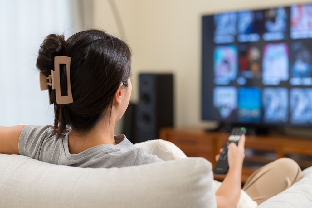Woman on couch watching TV, holding remote. Screen shows streaming service interface.