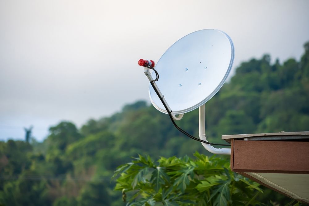 Satellite dish on a rooftop, with a green, forested backdrop and a cloudy sky.