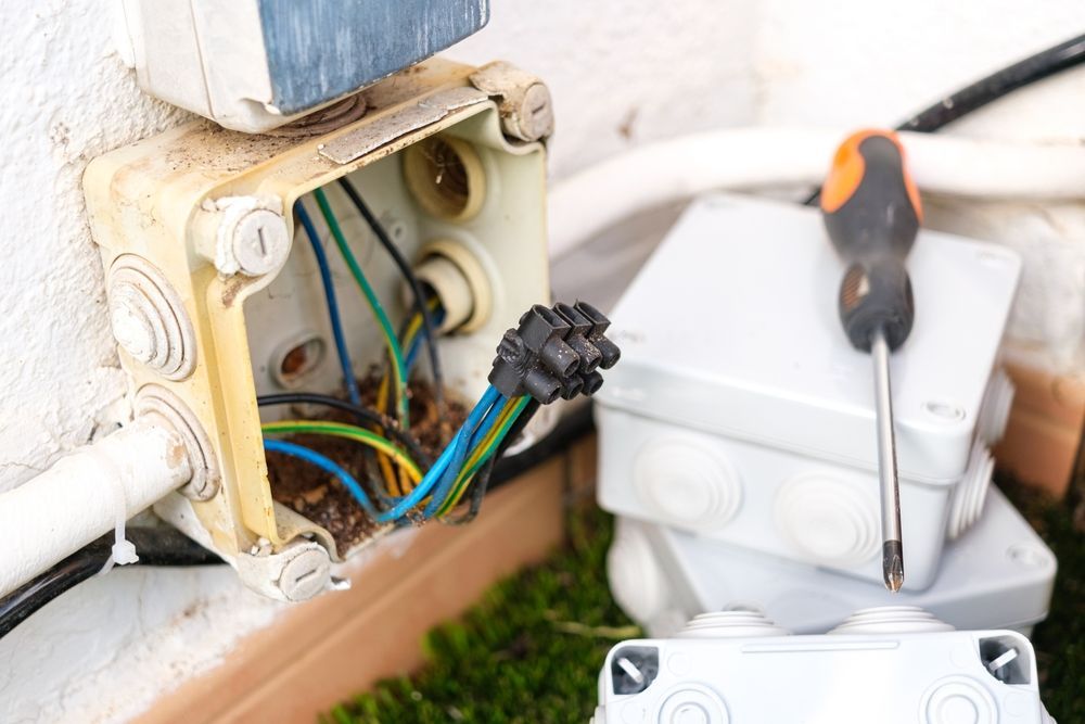 Damaged outdoor electrical junction box with exposed wires, screwdriver, and spare boxes.