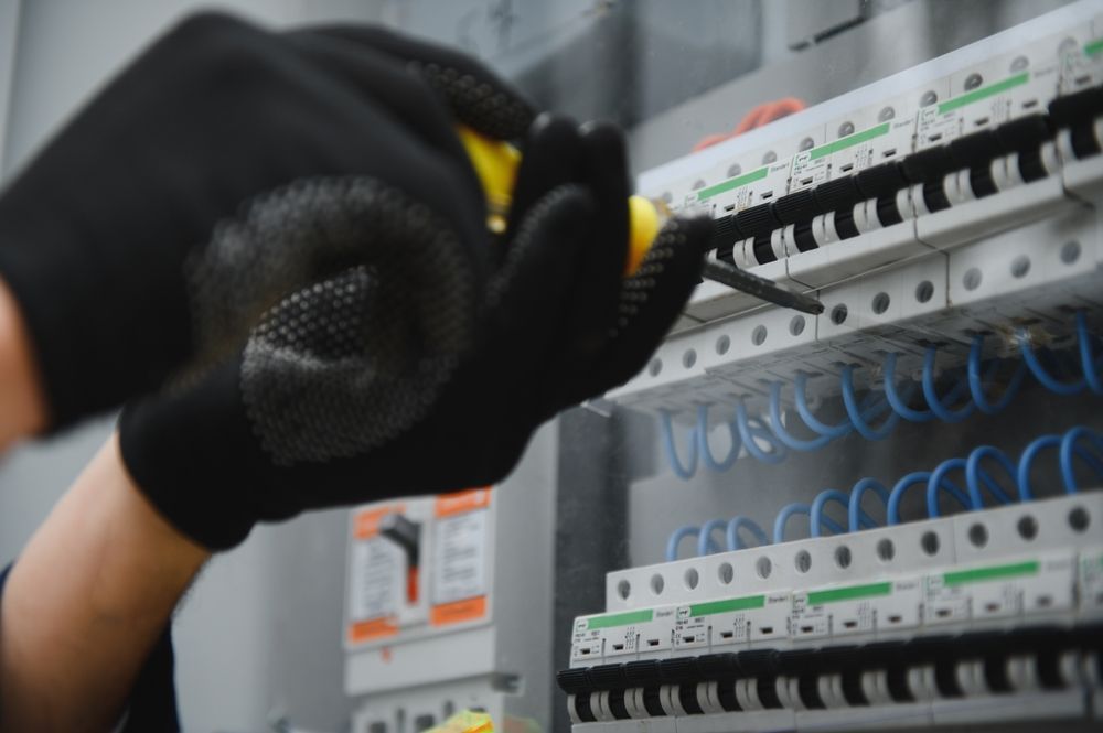 A person wearing black gloves using a yellow-handled screwdriver to work on a circuit breaker panel.