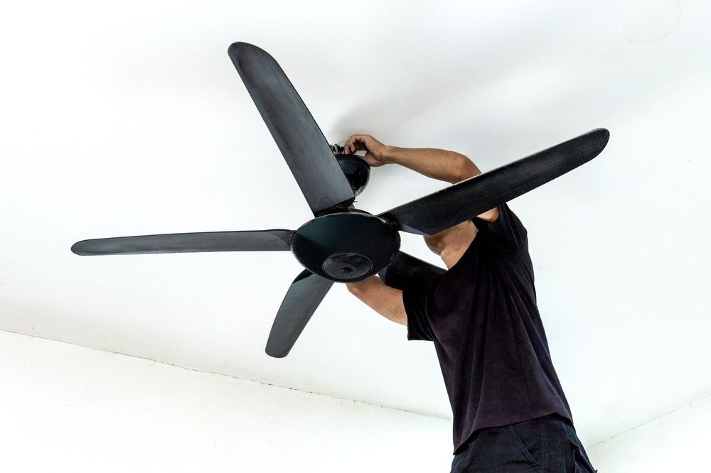 Person installing a black ceiling fan on a white ceiling.