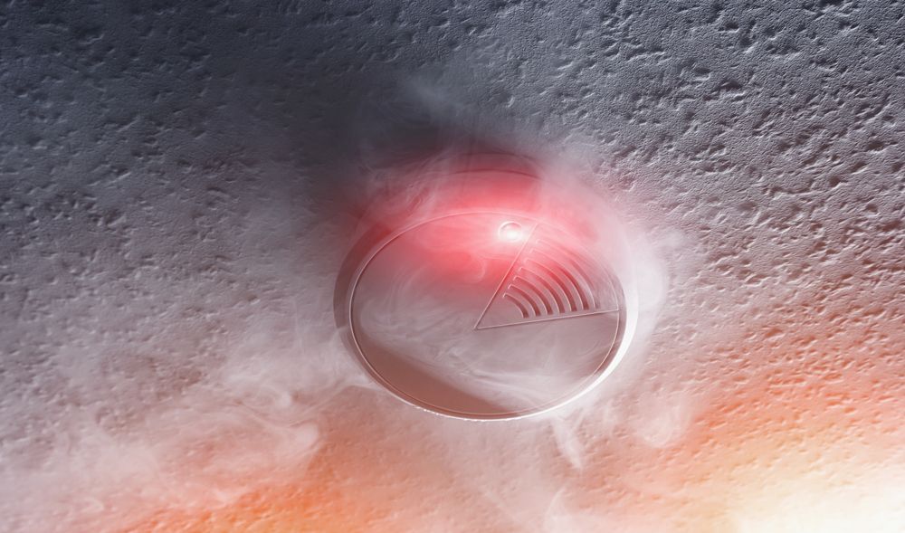Smoke detector with red light activated, surrounded by smoke on a textured ceiling.