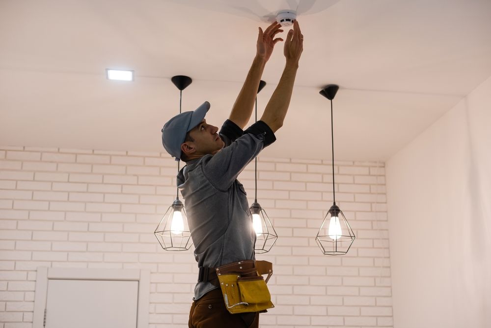 A person installs a smoke detector on a ceiling. They stand with arms raised, in a room with light fixtures.