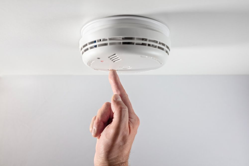 Hand pressing the test button of a white smoke detector on a white ceiling.