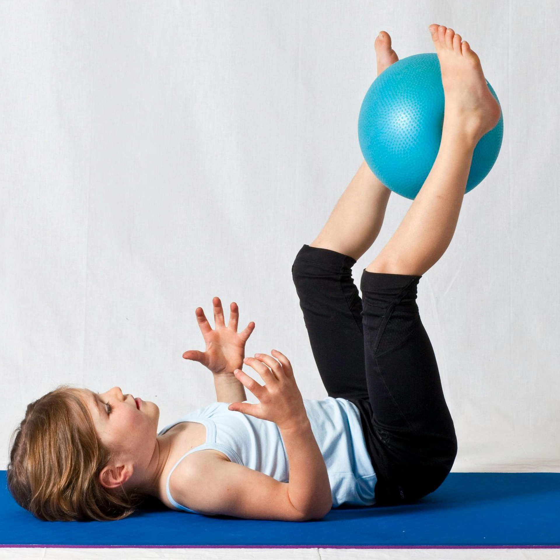 Young girl lying on a blue mat, holding a blue exercise ball between her feet, arms reaching out.