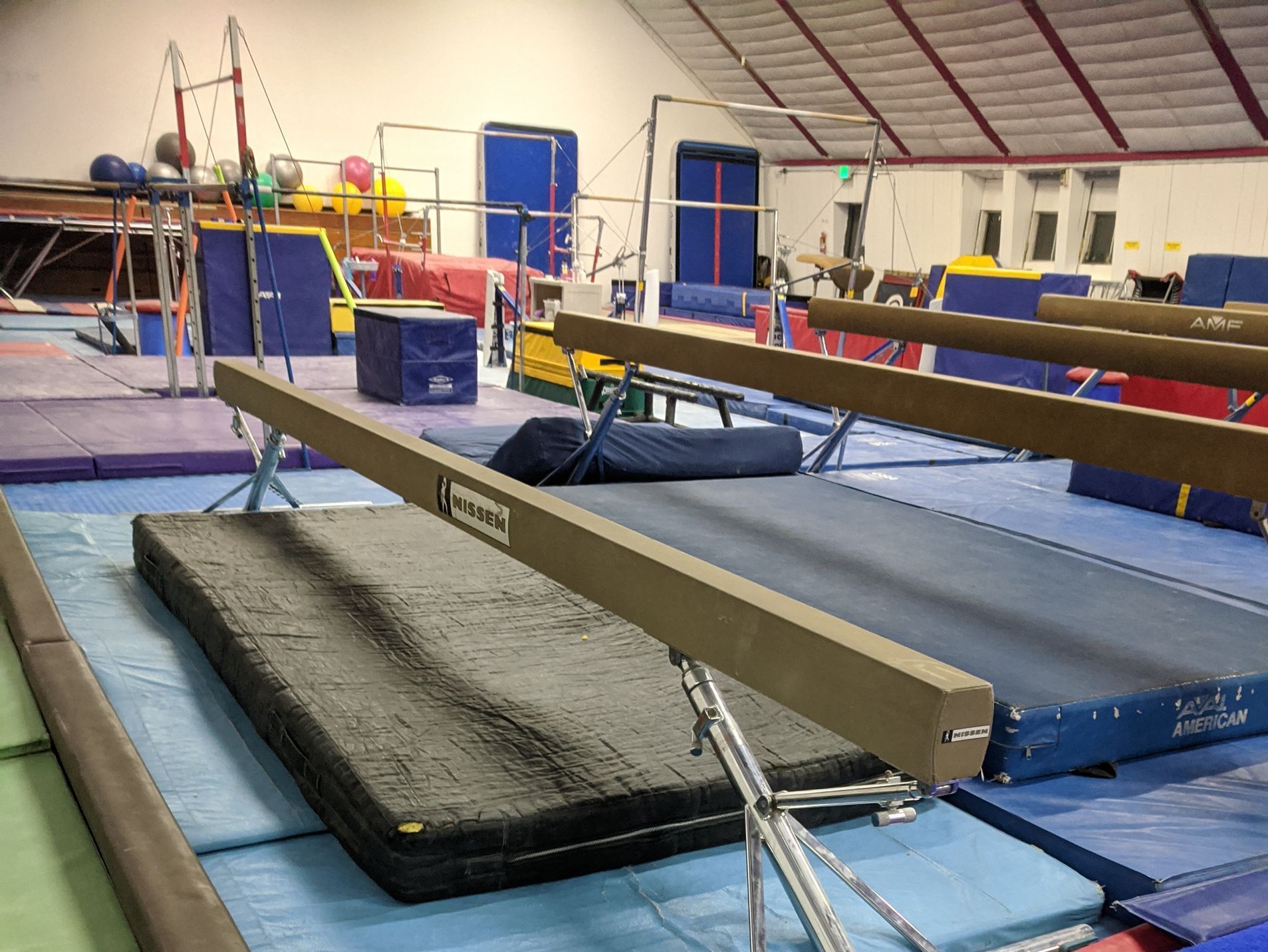 Gymnastics beams on blue mats in a gym setting. Other equipment like bars and mats are visible in the background.
