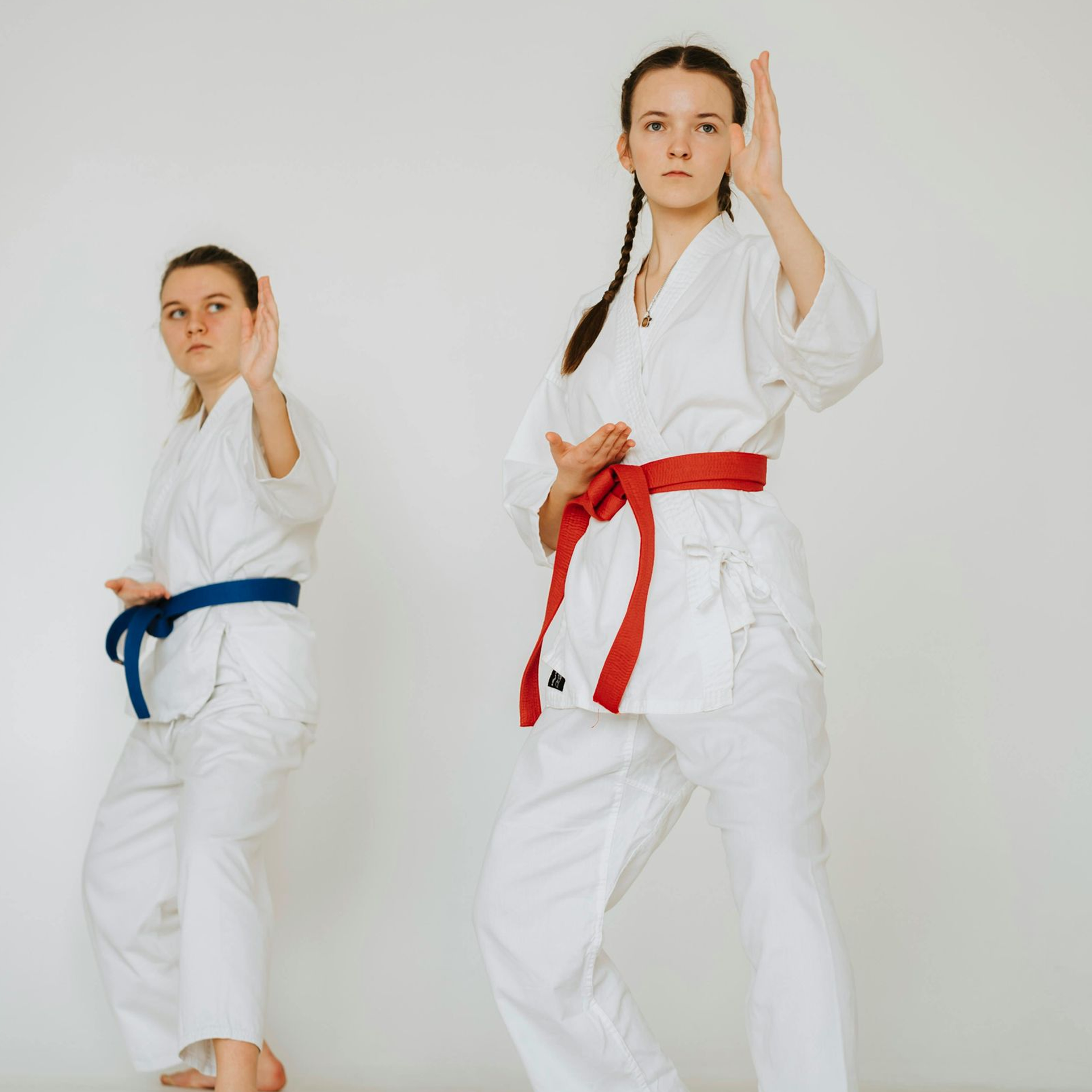 Two young women in white karate uniforms with colored belts practice a martial arts pose against a white background.