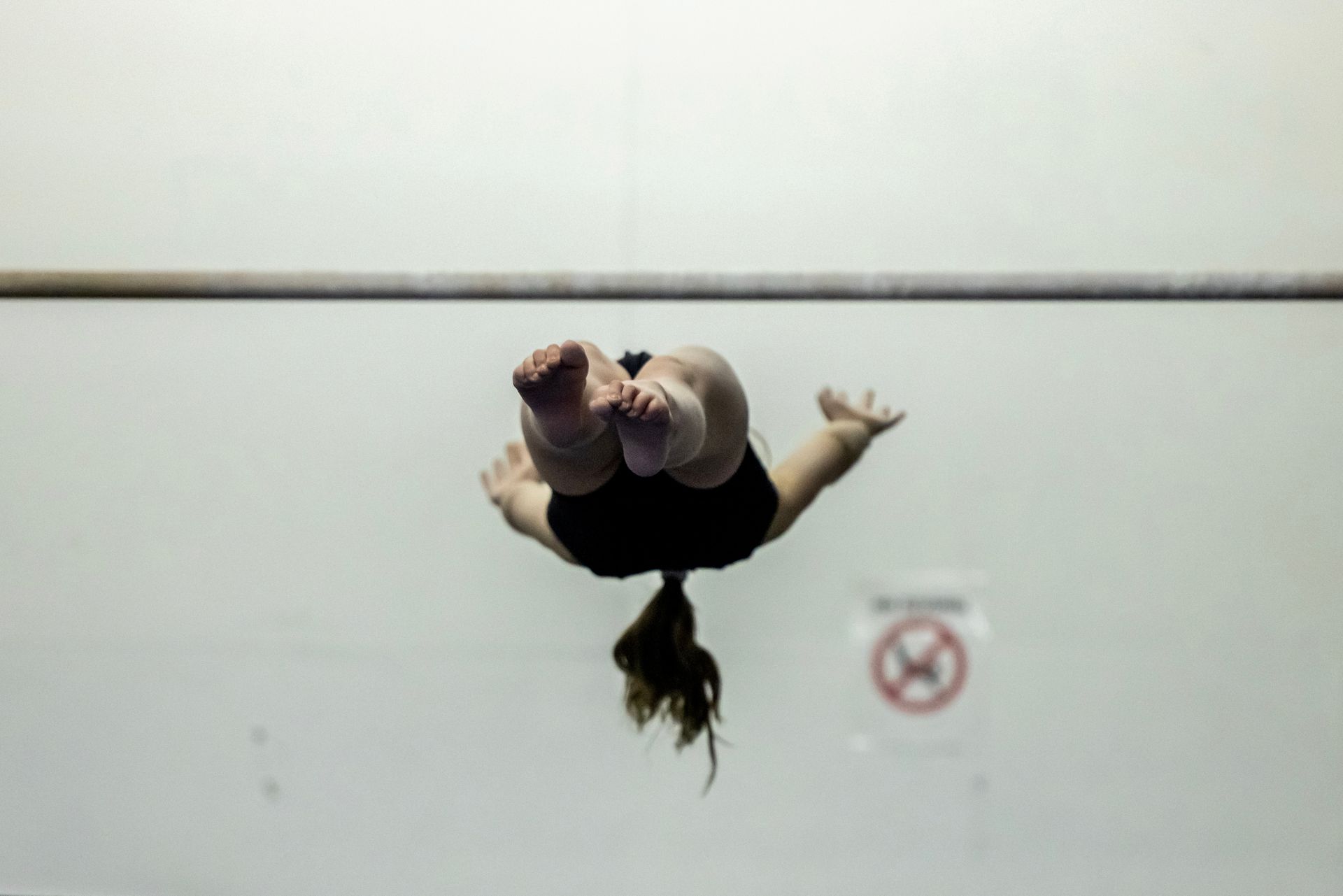 Gymnast performing a backflip above a bar, viewed from below, in a white room. She wears black and has her hair in a ponytail.
