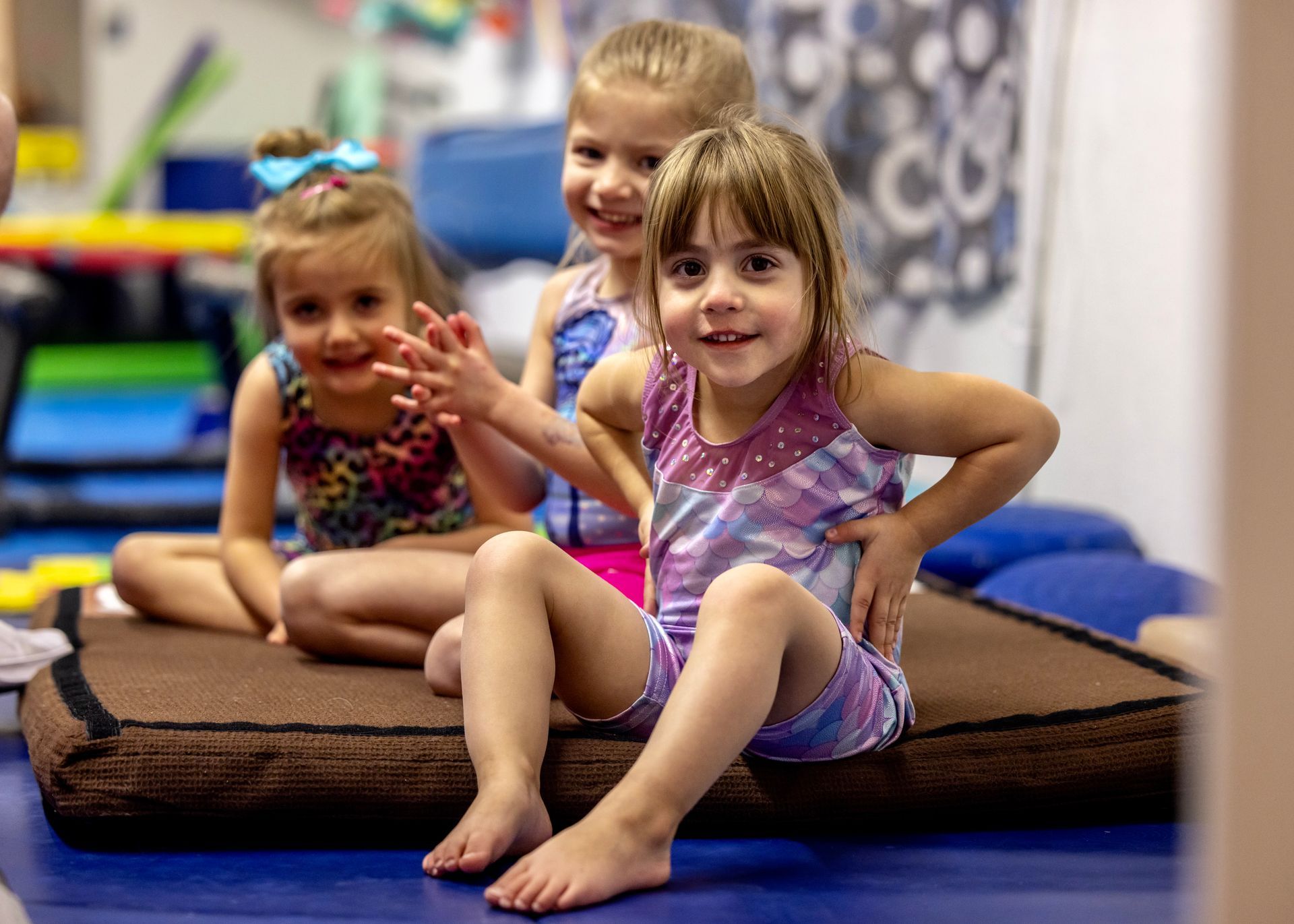 Three young girls in gymnastics attire smile while sitting on a brown mat at a gym.