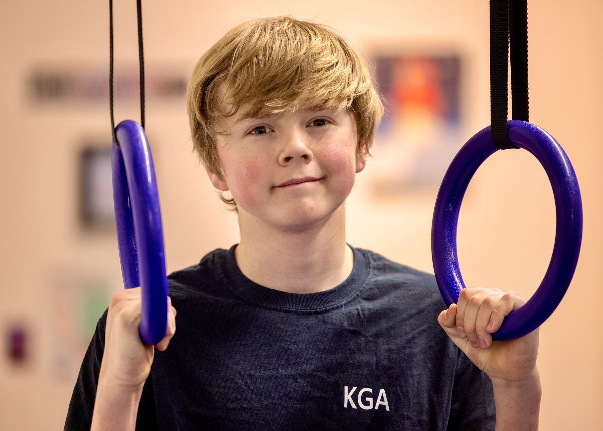 Young boy with reddish-blond hair smiling, holding gymnastic rings in a gym, wearing a navy blue shirt.