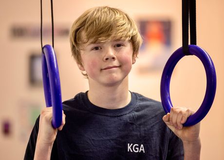 Young boy with reddish-blond hair smiling, holding gymnastic rings in a gym, wearing a navy blue shirt.