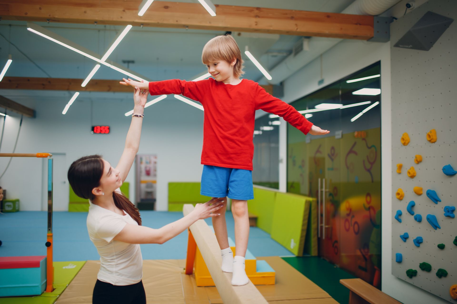 A woman assists a young boy balancing on a beam in a gym. The boy smiles, arms outstretched, as the woman steadies him.
