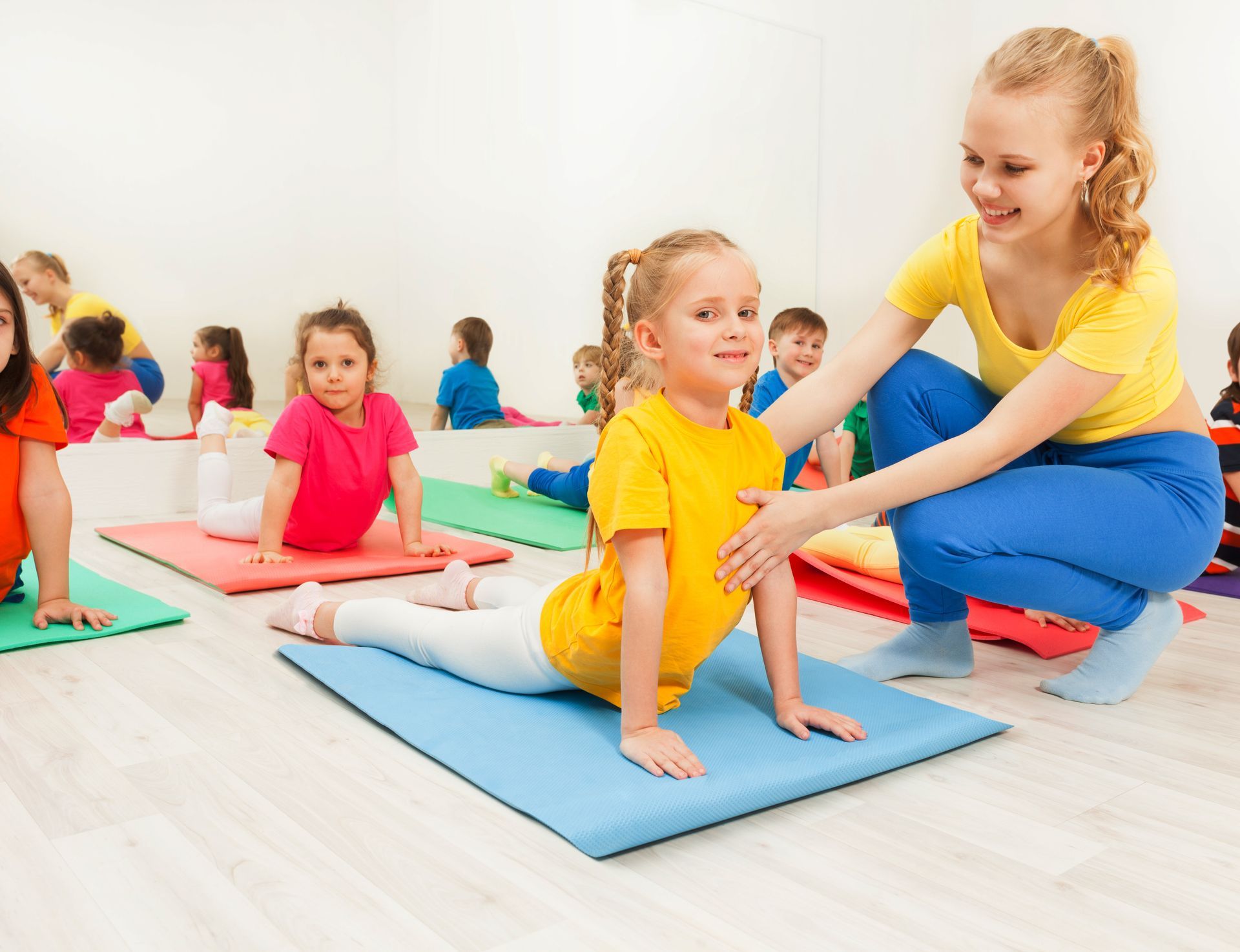 A young woman helps a girl in a yoga pose on a mat, in a room with other children.