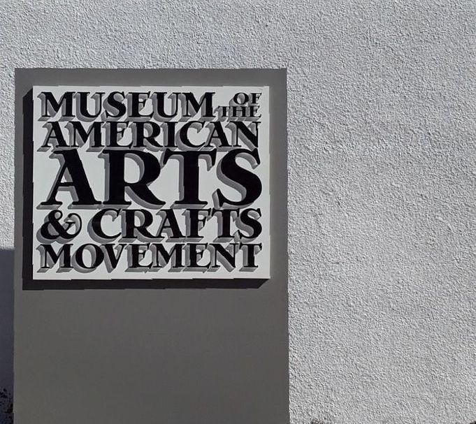 Sign for the Museum of the American Arts & Crafts Movement; black text on a light background, gray post, gray textured wall.