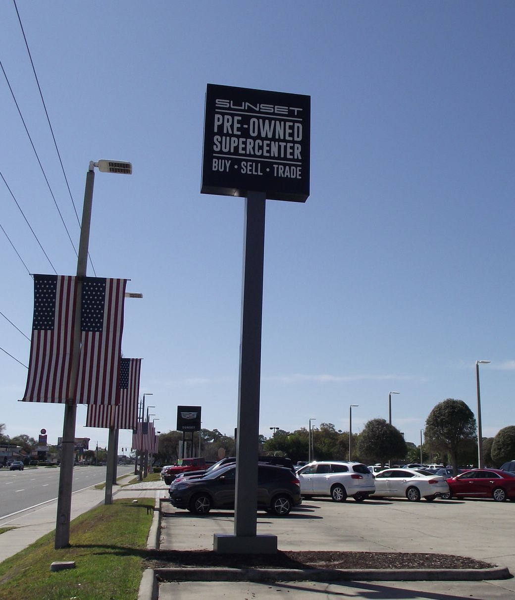 Sign for Sunset Pre-Owned Supercenter with US flags, parked cars, and a blue sky.