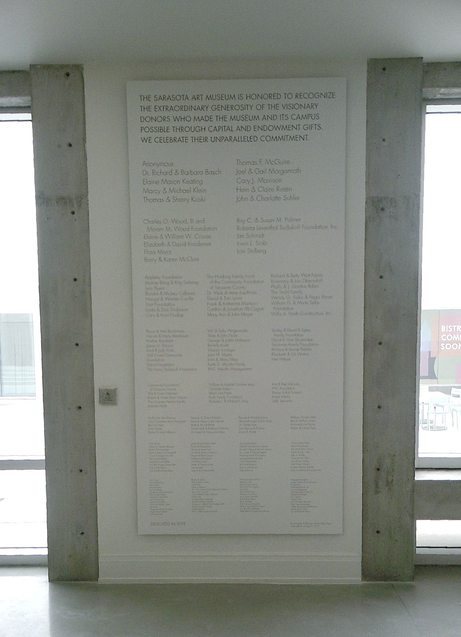 A white wall donors plaque with vinyl letters, set between two concrete pillars under construction, near a window.