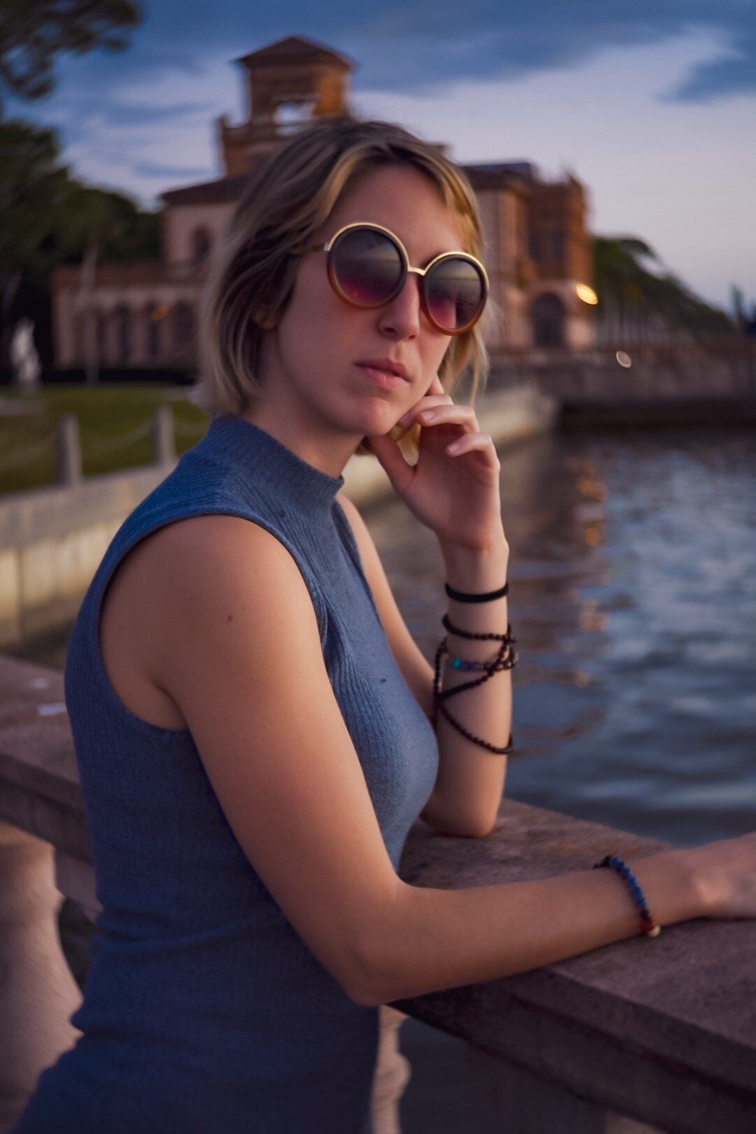 A woman wearing round sunglasses is leaning on a railing at the Ringling Museum overlooking a body of water.