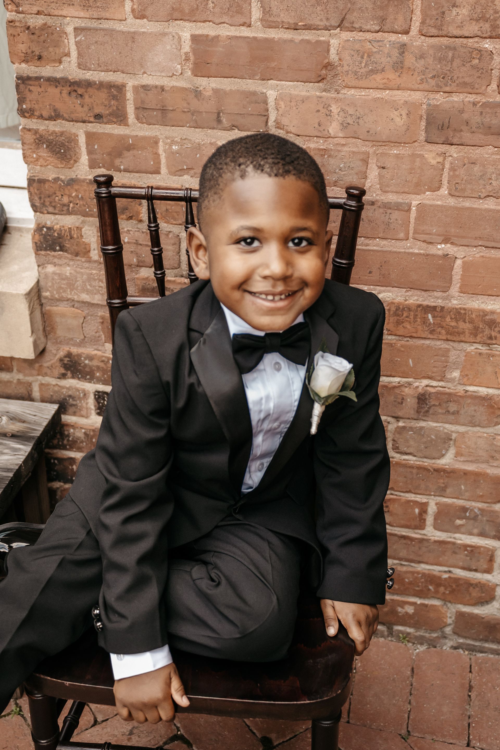 A young boy in a tuxedo is sitting on a chair in front of a brick wall.