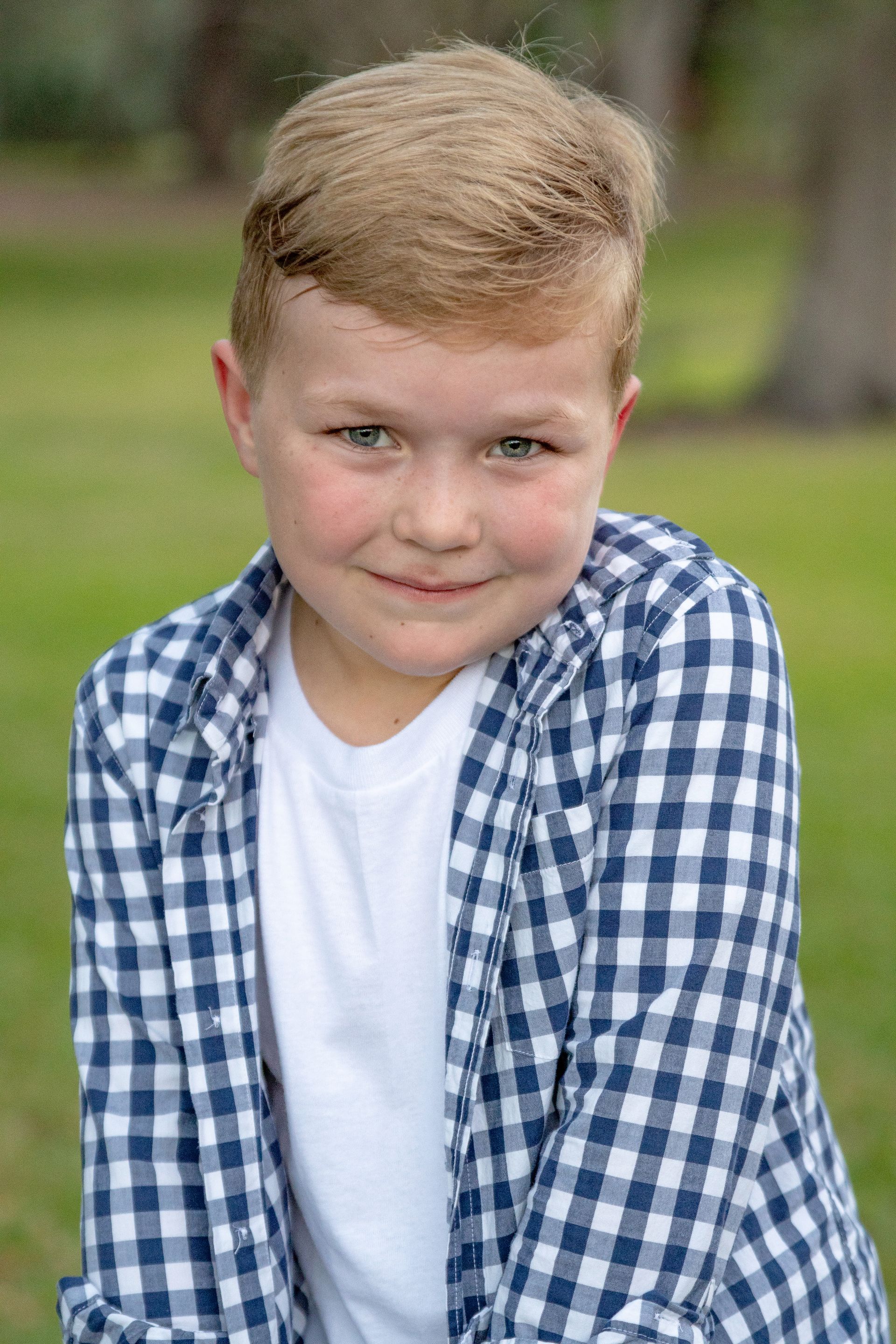 Blond boy in blue and white plaid shirt smiles outside.
