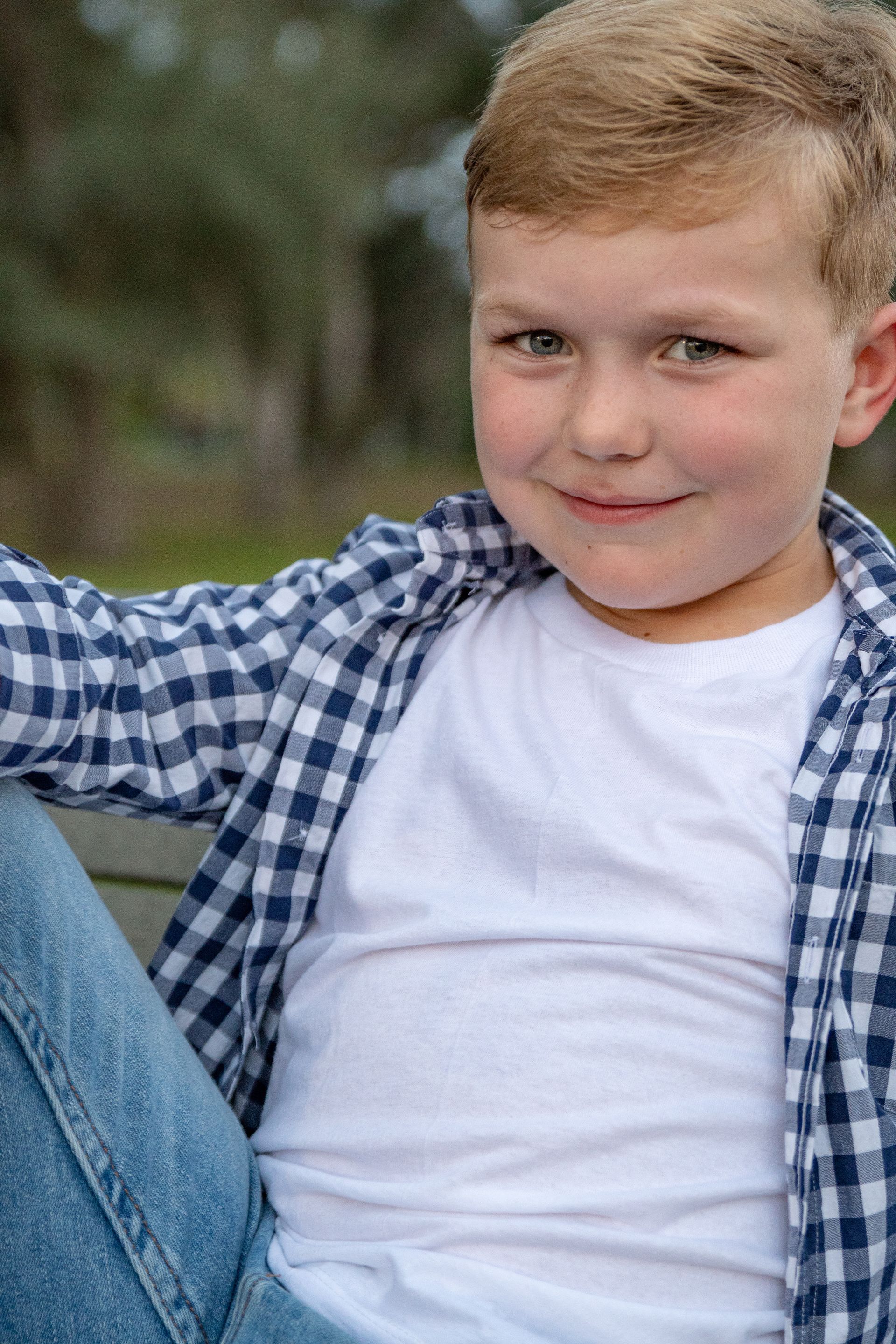 Young boy with blond hair, smiling, wearing a white shirt, blue jeans, and a blue checkered shirt.