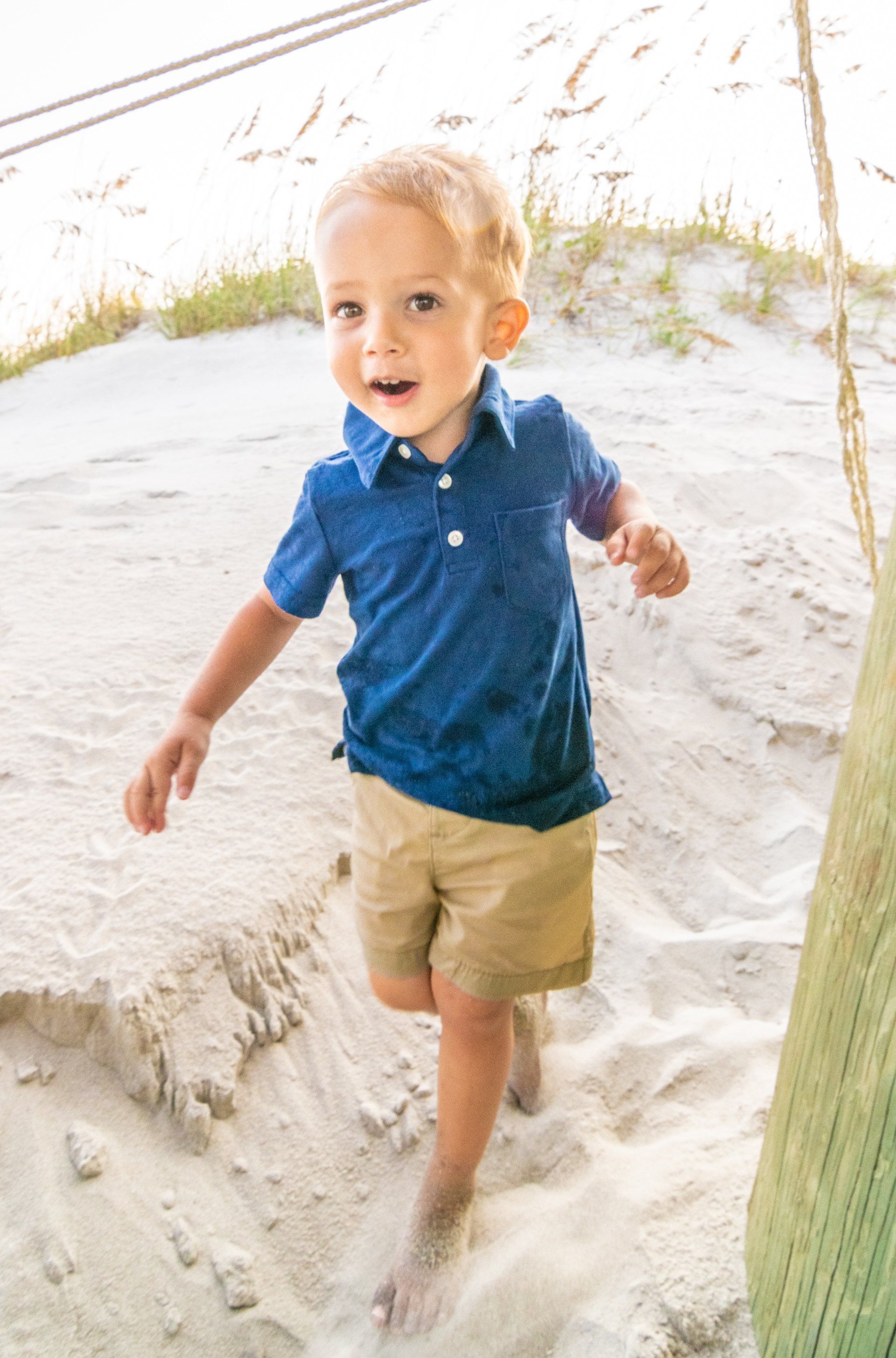 A little boy in a blue shirt and khaki shorts is standing in the sand on the beach.