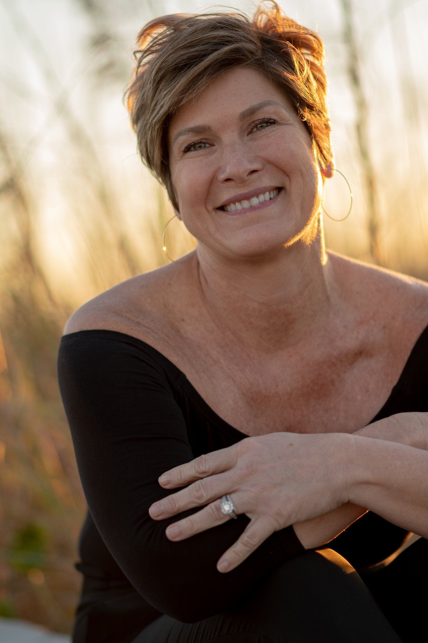 A woman wearing a black off the shoulder top and a ring on her finger is smiling.