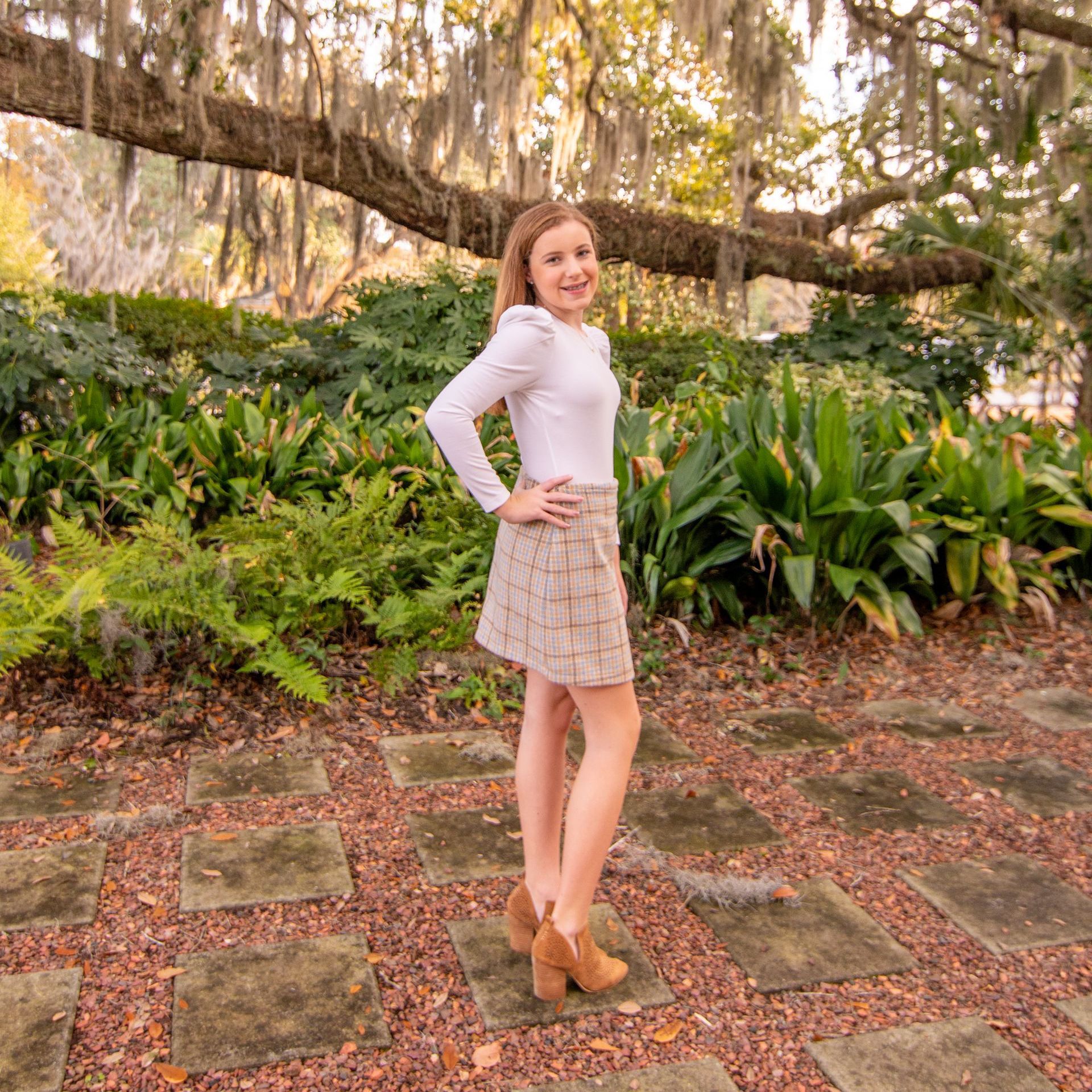 A woman in a white top and plaid skirt is standing in a garden.
