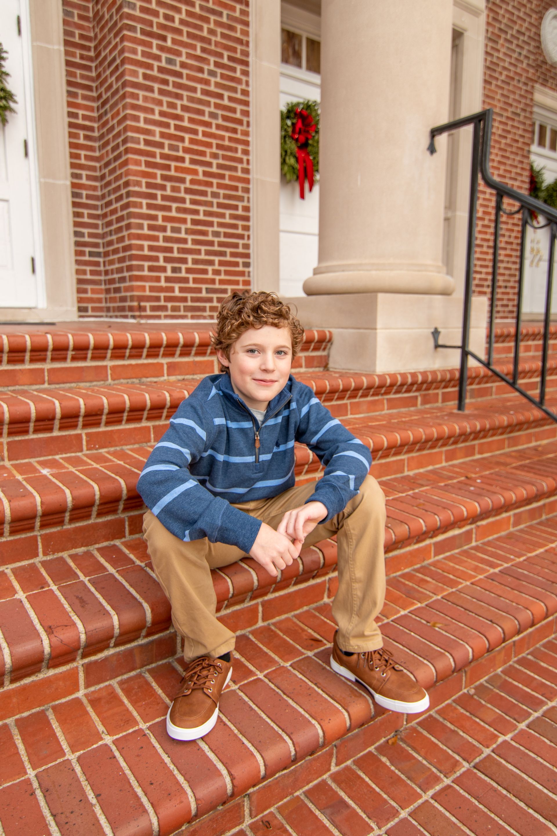 Boy with curly hair in blue shirt and khaki pants sits on brick steps.