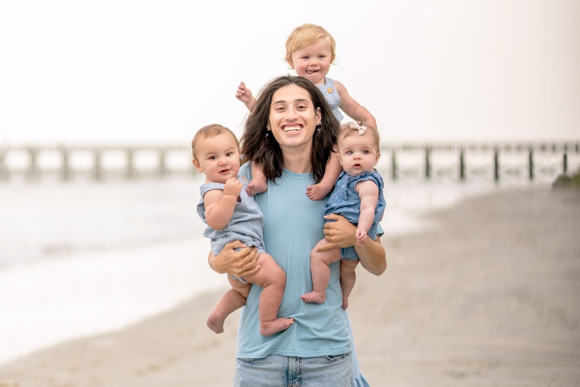 A man with long hair is carrying two babies on his side and a baby on his shoulders on the beach in North Myrtle Beach, SC.