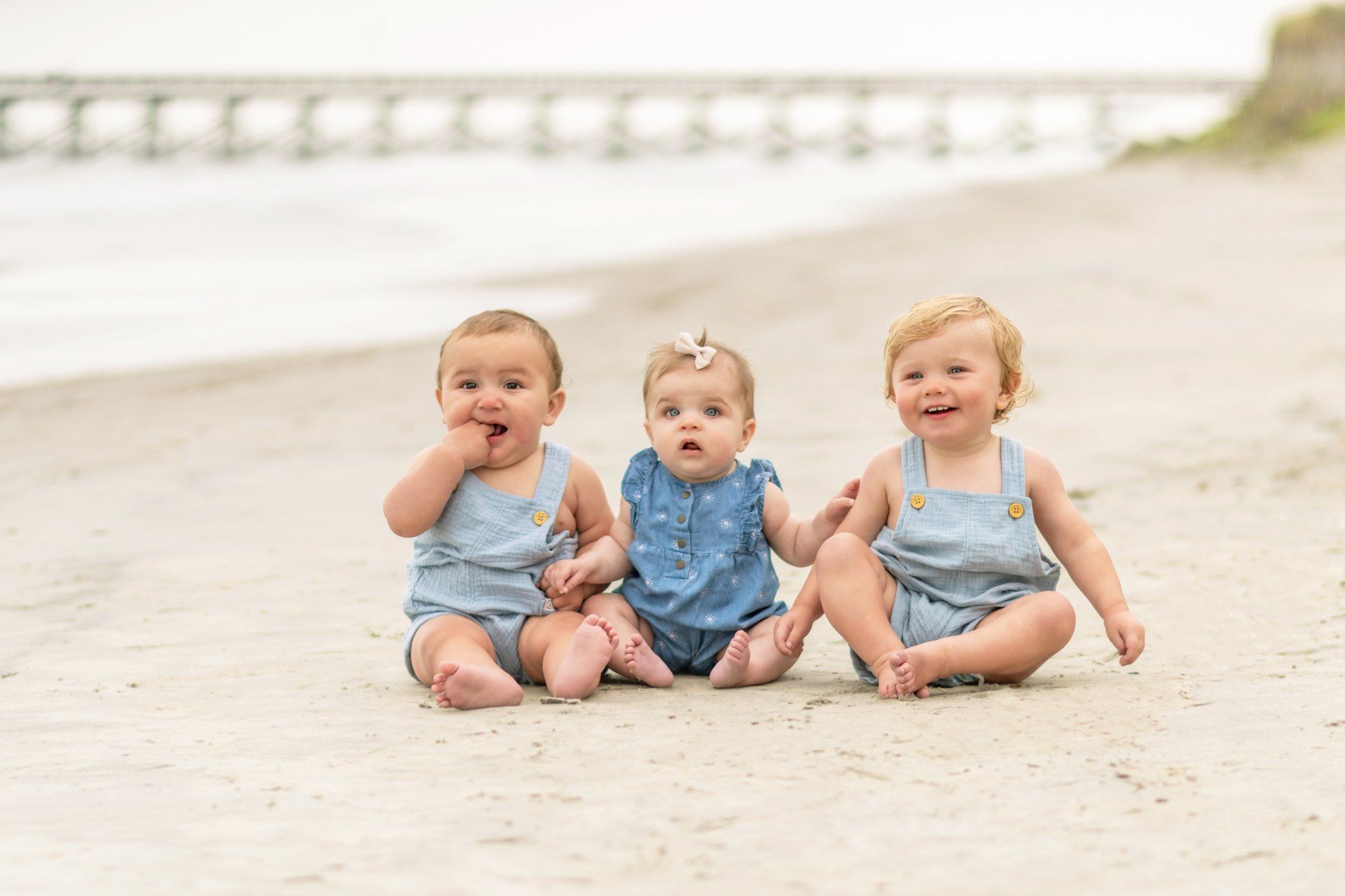 Three babies in blue are sitting on the beach together smiling and holding each other.