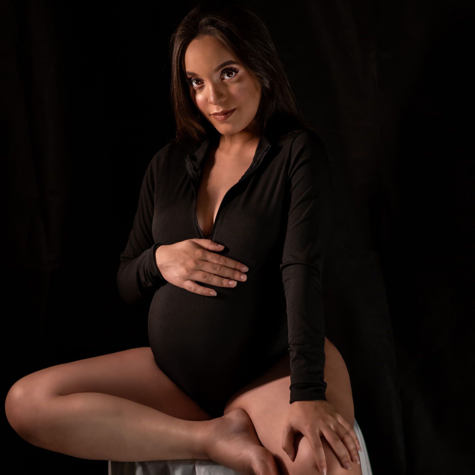 A pregnant woman in a black bodysuit is sitting on a chair holding her belly.