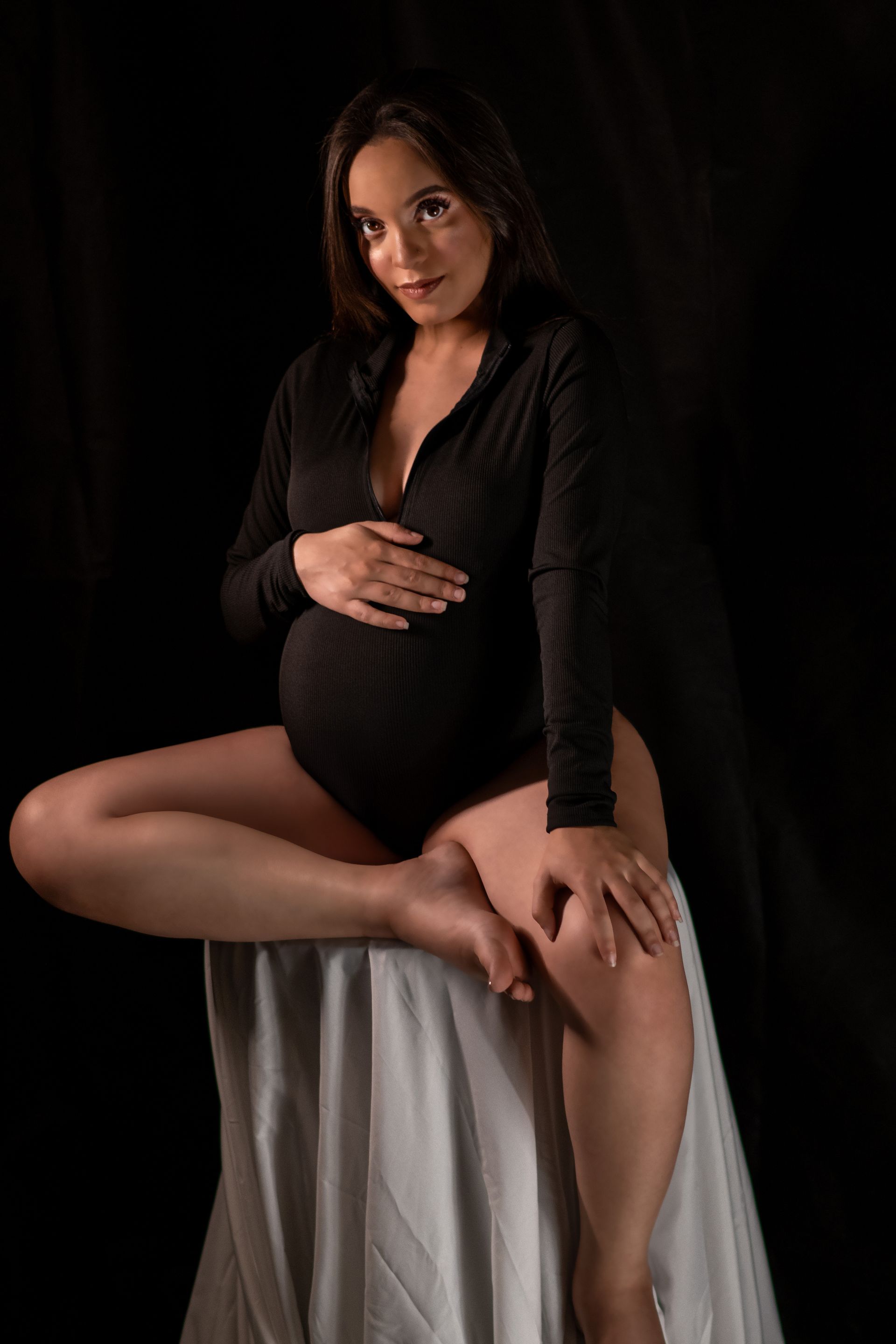 A pregnant woman in a black bodysuit is sitting on a table holding her belly.